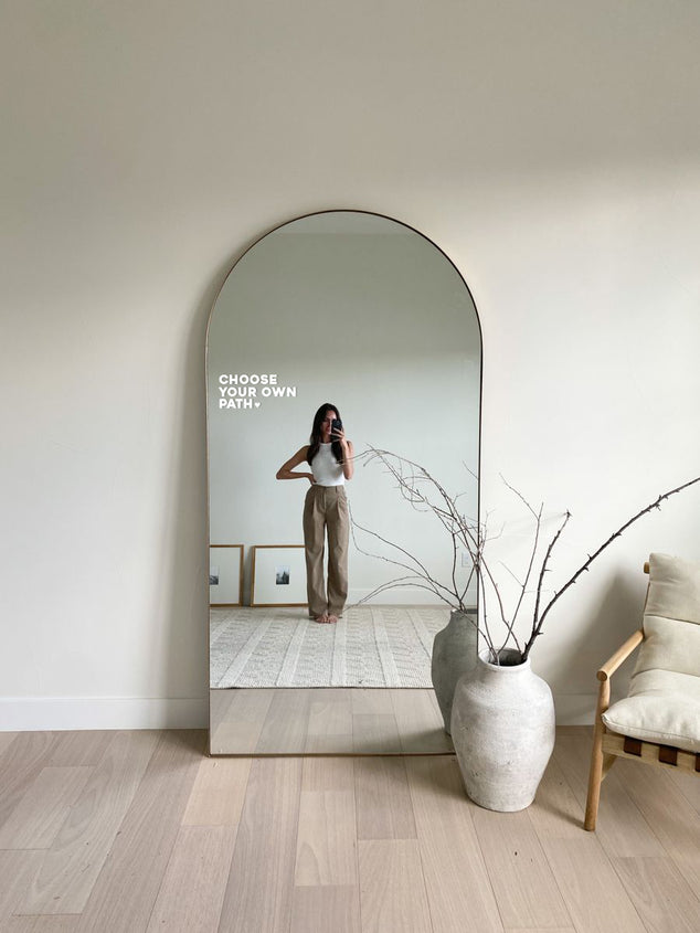 large freestanding mirror against loungeroom wall with white affirmation sticker 'choose your own path' and woman standing in mirror reflection