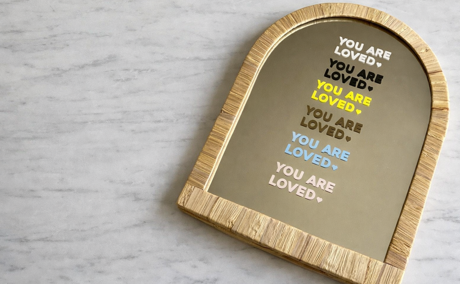 Decorative mirror with 'You are loved'  text shown in the 6 colours available for affirmation stickers on a marble table top